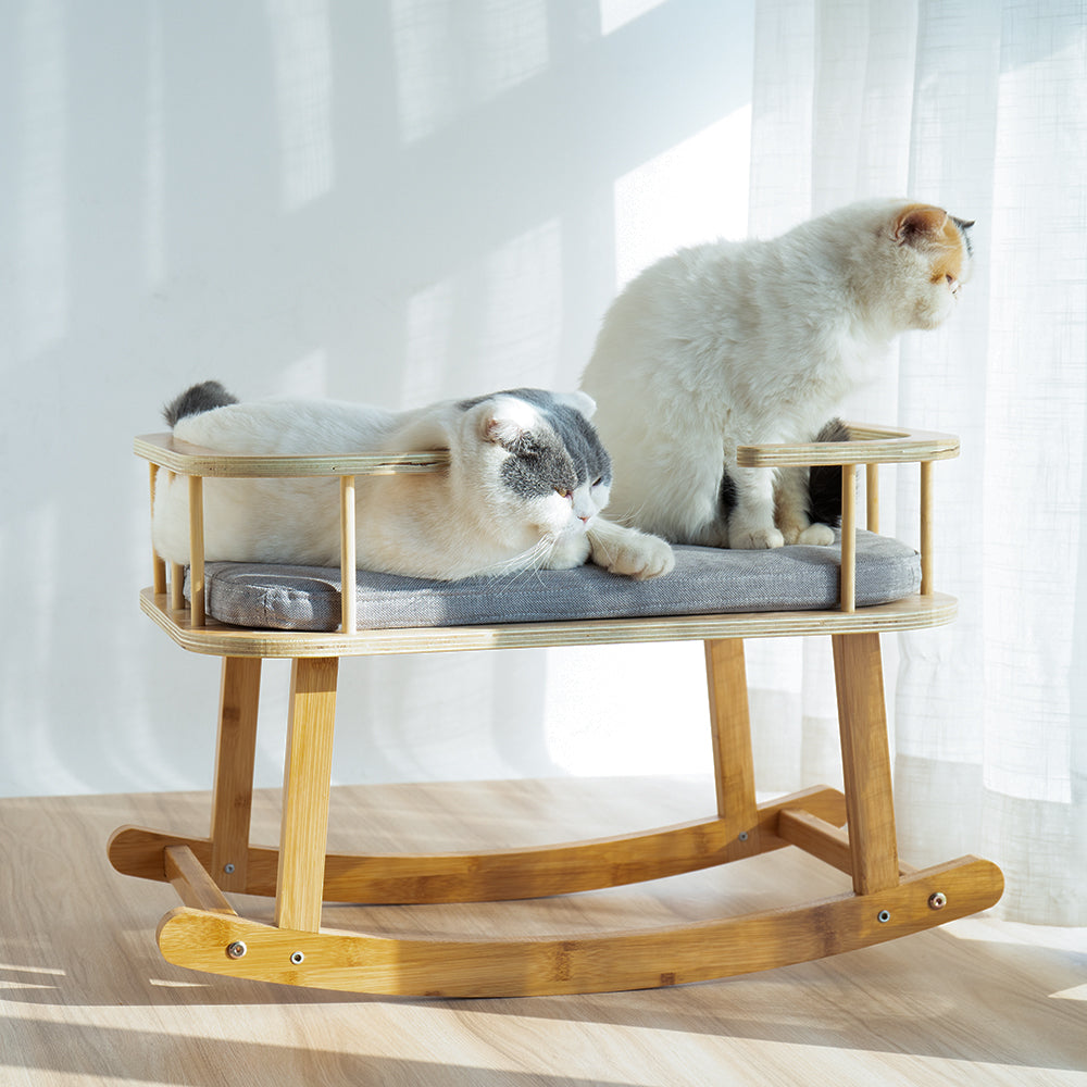 Two cats on a wooden rocking cat bed with a soft cushion in a bright room.